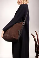 Bolso tipo bucket de gamuza marrón, con detalles y correa ajustable de cuero liso en tono más oscuro, hebilla metálica tipo estribo y costuras pespunteadas a la vista.