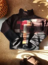 Buzo de lana uruguaya con diseño patchwork geométrico inspirado en el Tetris. Presenta una combinación de texturas y colores en tonos marrón, negro, rojo y gris, con una etiqueta de cuero en la parte inferior trasera.