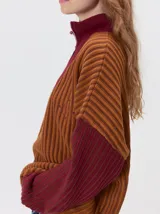 Pullover de tejido acanalado grueso con cuello alto y cierre frontal parcial. Presenta un diseño de bloques de color, con cuerpo en tono beige y mangas en contraste con rayas verticales en tonos rojizos. Incluye un detalle bordado en el pecho.