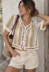 Blusa corta de tejido abierto tipo crochet, con mangas cortas, escote en V y cierre frontal con botones. Cuenta con bordados florales coloridos a lo largo de la botonera y el ruedo.
