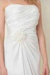 Vestido largo blanco de satén de seda natural, con breteles finos, escote recto, drapeado en el busto, tajo lateral y cola corta. Presenta aplique de flor de organza bordada en un costado.