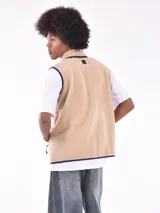 Chaleco de polar tipo corderito en color beige, con cuello alto y cierre frontal completo. Presenta un bolsillo de parche en el pecho en color azul marino con cierre de cremallera y ribetes en contraste en el mismo tono en sisas, cuello y bolsillos laterales.