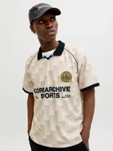 Camiseta tipo polo de manga corta con calce suelto. Presenta un diseño de cuadros en tonos beige y crema, con cuello y bordes de mangas en color negro. Incluye un escudo circular bordado en el pecho y estampado tipográfico en el frente.
