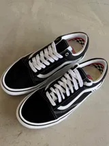Championes Vans Skate Old Skool, color negro con franja lateral blanca, cordones blancos y suela blanca con línea negra. Presentan un detalle de bandera a cuadros en el lateral.
