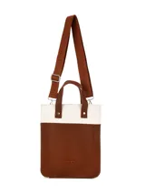 Matera de diseño vertical con combinación de materiales en color beige y marrón. Presenta cuerpo inferior en cuero sintético marrón con logo grabado y sección superior en tejido de lona cruda. Incluye asas cortas de mano en cuero sintético marrón con remaches metálicos.