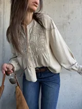Camisa beige de manga larga con detalles bordados en el frente, cuello redondo y cierre frontal con botones. Las mangas presentan puños con volados.