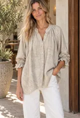 Camisa amplia de tejido liviano color beige, con escote en V y cuello mao. Presenta frunces en los hombros, cierre frontal con botones y mangas largas terminadas en puños elastizados con volados.