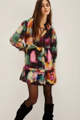 Vestido corto con estampado abstracto en tonos vibrantes como fucsia, verde, amarillo y negro. Presenta cuello con lazo ajustable y mangas largas abullonadas con puños elásticos. El cuerpo tiene un corte imperio con un volante fruncido en el ruedo.