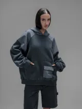 Hoodie oversize de color gris oscuro, confeccionado en frisa de algodón con recortes en textiles técnicos. Presenta capucha forrada, bolsillo frontal tipo canguro y un bolsillo adicional con solapa en la parte inferior.