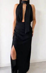 Vestido largo negro con escote halter profundo y detalle de lazo al cuello. Presenta un diseño drapeado en la cintura y una abertura lateral pronunciada en la falda.
