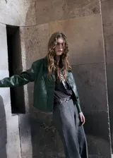 Chaqueta corta de efecto piel en color verde oscuro, con diseño recto, cuello camisero, manga larga y cierre frontal de botones ocultos. Presenta dos bolsillos de parche con solapa en la parte delantera y cuenta con forro interior.