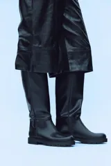 Bota plana de caña alta en color negro, con diseño minimalista y detalle de pespuntes verticales en el cuerpo. Cuenta con suela track, punta redonda y tirador trasero para facilitar el calce.