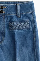 Pantalón de jean estilo wide leg con tiro alto, confeccionado en denim de algodón rígido. Presenta bolsillos delanteros con detalle de ribete trenzado, pliegues frontales y traseros, y cierre con doble botón.