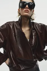 Chaqueta de corte amplio estilo biker en tejido efecto piel de color verde oscuro, con cuello alto envolvente, cierre frontal asimétrico de cremallera metálica, bolsillos ocultos en costura y forro interior.
