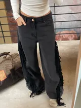 Pantalón de jean negro de corte amplio y tiro medio-alto. Presenta flecos laterales de cuero genuino a lo largo de las piernas.