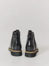 Bota corta de cuero negro con textura de reptil, diseño tipo chelsea con elásticos laterales y costuras a la vista en tono contrastante. Cuenta con suela plana de goma con detalle de franja en color natural.