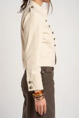 Chaqueta corta color beige de algodón, de inspiración militar con cuello mao y doble botonadura frontal con botones metálicos dorados.