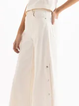 Pantalón de denim de algodón pima color blanco off-white, de tiro alto y silueta amplia de corte ancho. Presenta costuras verticales visibles y broches metálicos decorativos y funcionales en los laterales.