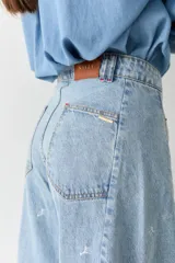Jean de corte wide leg y tiro alto, confeccionado en denim de lavado claro. Presenta un diseño con costuras verticales frontales y un sutil estampado de figuras deportivas pequeñas distribuidas por toda la prenda.
