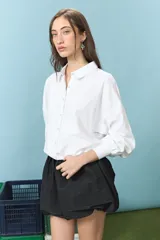 Camisa blanca de corte oversize con cuello camisero y mangas largas. Presenta un bordado en la espalda con el diseño de un escudo nacional estilizado en color azul y la frase 'El país de los mates y las siestas'.
