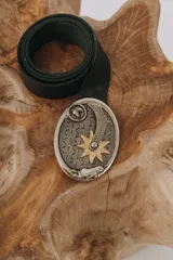 Cinturón negro de cuero con hebilla ovalada plateada y dorada con motivos country: cabeza de caballo, pluma, estrella y lagartos.