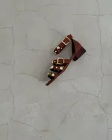 Sandalia de cuero marrón con tiras finas y detalles de tachas doradas. Tiene un pequeño taco cuadrado y cierre con hebilla dorada en el tobillo.