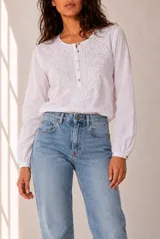 Blusa blanca de manga larga con detalles bordados en tono marrón en el pecho y las mangas, cuello redondo y cierre frontal con pequeños botones.