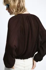 Chaqueta de organza plisada color marrón oscuro, con cuello redondo, cierre frontal con cremallera metálica, mangas largas con puños elásticos y bajo con acabado elástico.