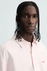 Camisa de algodón color rosa pálido, corte boxy fit, con cuello solapa, manga corta y abotonadura frontal. Presenta un texto bordado en contraste de color rojo en el delantero.