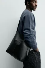 Bolso tipo shopper de cuero sintético negro, con costuras visibles en el frente que dividen el cuerpo en paneles. Presenta un borde superior ondulado y un asa de hombro ajustable.