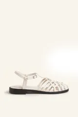 Sandalia plana de cuero blanco con diseño de tiras trenzadas en la parte delantera. Cuenta con una tira ajustable al tobillo con hebilla metálica y suela plana de color negro.