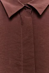 Camisa de cuello solapa y manga corta con dobladillo. Presenta un diseño de corte holgado con bajo asimétrico y aberturas laterales. Cierre frontal mediante botones ocultos bajo una tapeta.