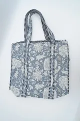 Bolso tipo tote bag de tela acolchada con estampado floral en tonos gris y blanco. Presenta asas largas reforzadas con detalle de rayas en los bordes.