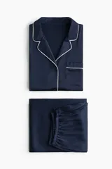 Pijama de dos piezas en satén azul marino con ribetes blancos en contraste. Camisa de manga larga con cuello solapa, cierre frontal de botones y bolsillo en el pecho. Pantalón de corte recto y pierna amplia con cintura elástica y cordón ajustable.