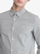Camisa de vestir gris claro de manga larga, con cuello clásico y cierre de botones. Tiene el logo de Calvin Klein bordado en el pecho.