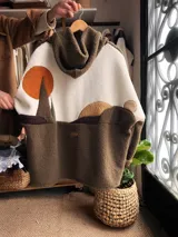 Campera tipo buzo con capucha confeccionada en tela sherpa, con diseño artesanal en la espalda que representa un paisaje de campo con sol y luna mediante apliques de tela. Presenta una combinación de colores verde oscuro y crudo, con cierre frontal y corte holgado.