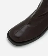 Bota tipo sock boot de caña media, confeccionada en poliuretano color marrón oscuro con acabado brillante. Presenta puntera redondeada, cierre lateral y taco bloque bajo de color negro.