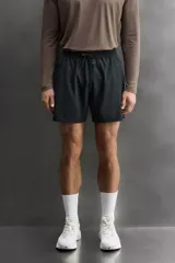 Pantalón corto deportivo color marrón oliva, confeccionado en tejido técnico ligero y elástico, con forro interior. Presenta cintura elástica ajustable con cordón, bolsillos frontales, bolsillo de seguridad trasero con cierre y aberturas laterales en el bajo.