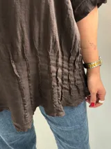 Blusa de lino color marrón chocolate, de corte holgado, con cuello redondo, mangas tres cuartos con trabilla y botón para sujetar remangadas, y detalle de alforzas verticales en el frente.