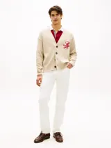 Cárdigan de punto grueso en color beige, con cuello en V, cierre frontal con botones y bolsillos de ojal. Presenta puños y bajo acanalados con líneas rojas y blancas, y un logo script bordado en rojo en el pecho.