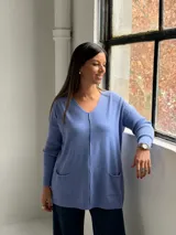 Sweater de punto color azul lavanda, con escote en V, mangas largas con puños acanalados y dos bolsillos frontales tipo parche. Presenta una costura central vertical visible.