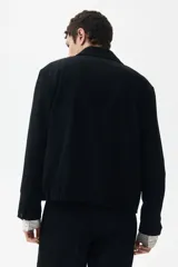 Campera negra de tejido suave, corte estándar, con cuello clásico, cierre frontal y bolsillos laterales con cierre. Presenta puños abotonados y elástico oculto en la parte posterior del bajo.