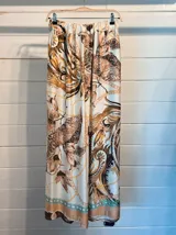 Pantalón palazzo de satén blanco con estampado barroco de hojas tropicales en tonos verde oliva y mostaza, y detalles de cadenas en color dorado y rosado. Presenta un borde inferior con detalles en tonos melocotón y azul claro.