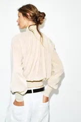 Blusa marrón de punto fluido, con cuello subido abotonado en un lateral y mangas largas con puños ajustados. Presenta un diseño fruncido en la parte frontal que aporta volumen.