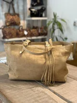 Cartera tipo tote de cuero gamuzado en color arena, con textura grabada tipo cocodrilo. Presenta doble asa con hebillas metálicas ajustables y detalle de flecos largos colgantes en uno de los laterales.