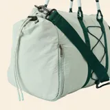 Bolsa de deporte color verde claro, con correas de hombro tipo cuerda, correa ajustable extraíble y sistema de cordones elásticos en el frente para sujeción.