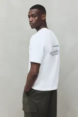 Remera blanca de algodón con cuello redondo y corte holgado. Presenta un estampado con texto en color negro en el frente.