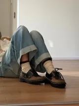 Mocasín de estilo náutico confeccionado en cuero, con diseño bicolor en negro y marrón. Presenta cordones, suela de goma dentada con costuras visibles y detalle de ojales metálicos.
