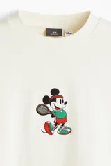 Buzo de tejido de algodón color crema, corte oversized y cuello redondo. Presenta un pequeño bordado central de Mickey Mouse vestido con ropa deportiva y sosteniendo una raqueta de tenis.