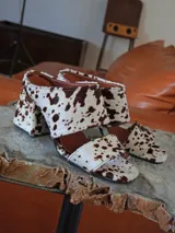 Sandalias de tacón alto y ancho, confeccionadas en piel con acabado de pelo y estampado animal print de vaca en tonos marrones y blancos, con tiras anchas en el empeine.